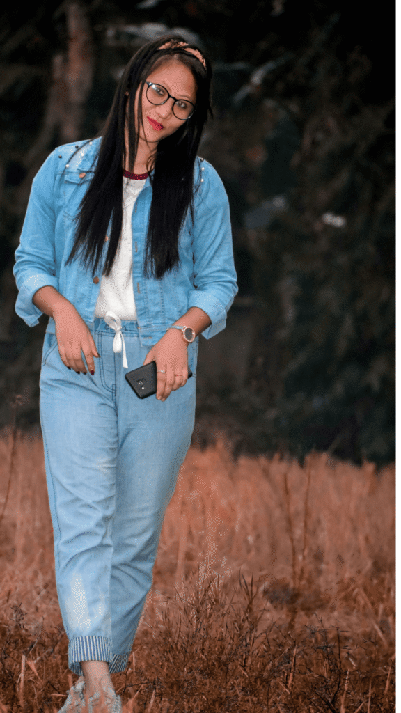 A woman with long dark hair and glasses posing outdoors in a light-wash denim outfit, wearing a light blue denim jacket over a white top with matching cuffed denim joggers featuring a drawstring waist.
