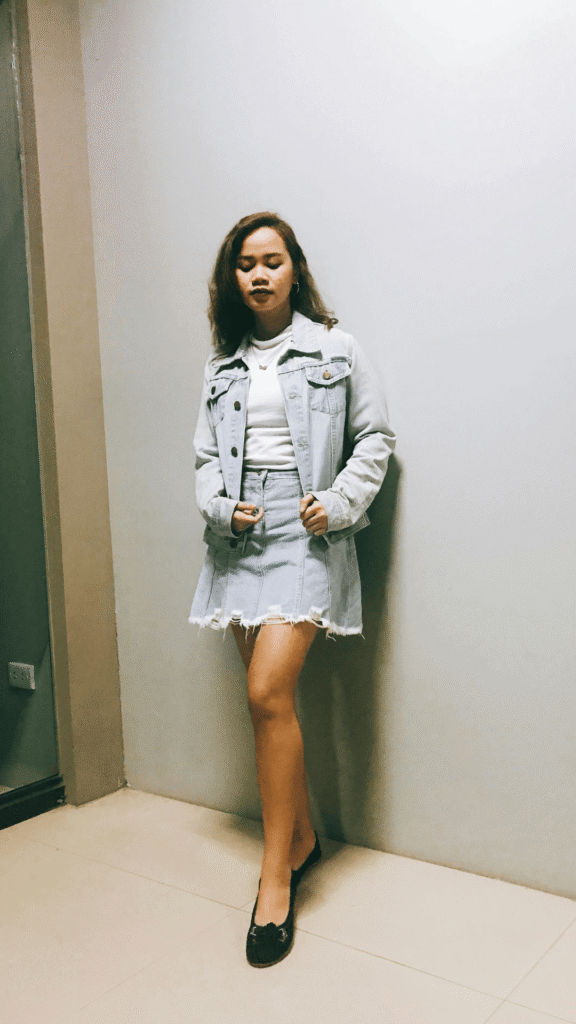 A woman posing indoors against a neutral wall wearing a light-wash denim jacket layered over a plain white high-neck top, paired with a matching light-wash distressed denim mini skirt with a frayed hem and dark flats.