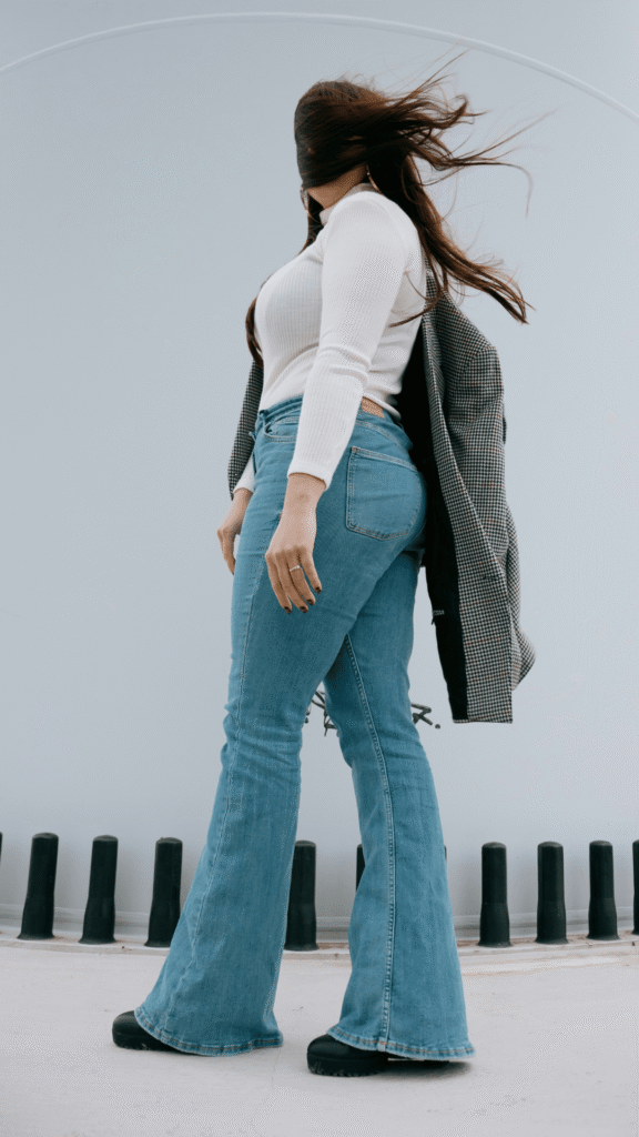 A woman with long brown hair blowing in the wind posing outdoors in high-waisted flared jeans, a fitted white ribbed long-sleeve turtleneck sweater, black chunky boots, and a houndstooth blazer draped over her shoulders.